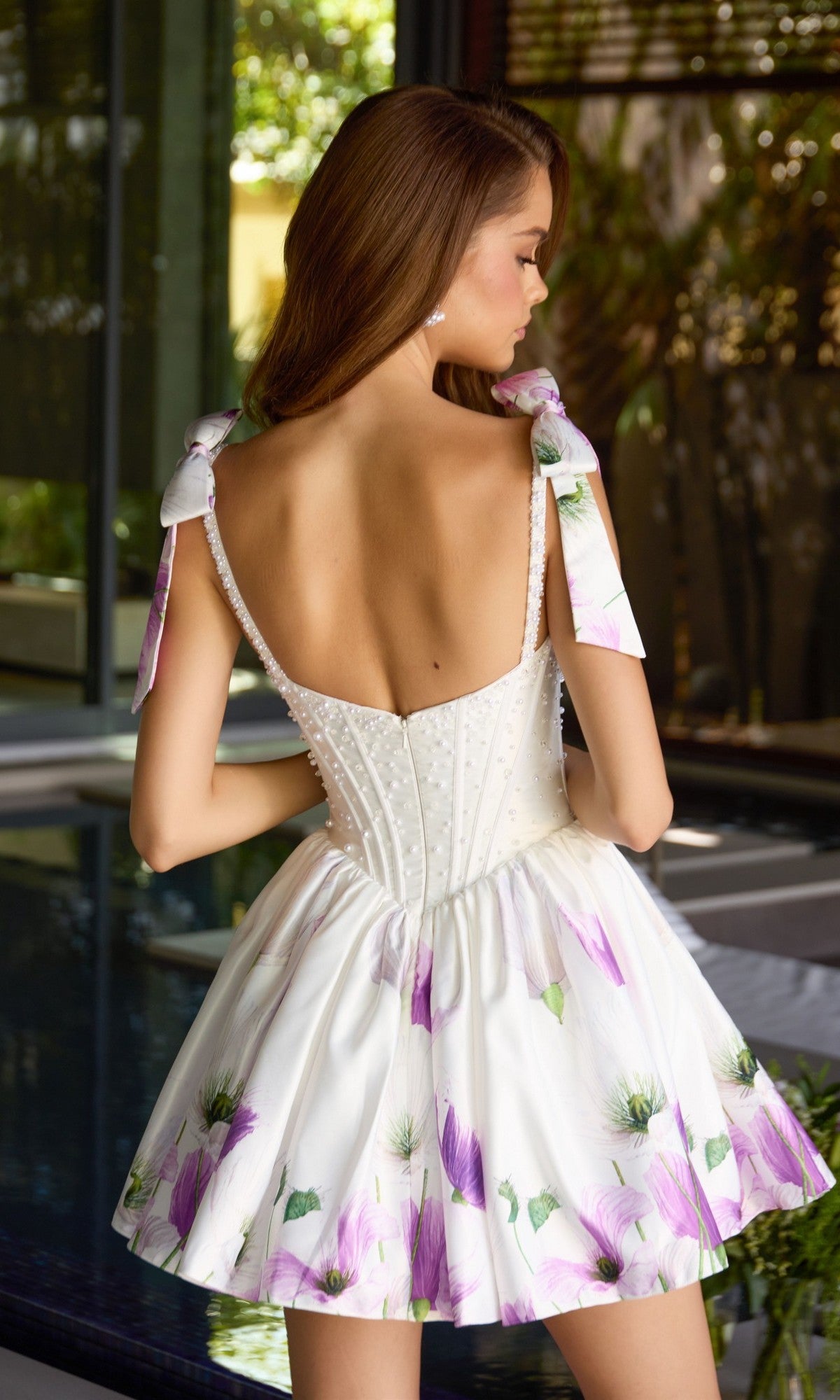 A woman faces away from the camera, modeling the Ava Presley 42432 short cocktail dress in floral satin, adorned with purple blooms, pearl accents, and bow-tied shoulder straps.